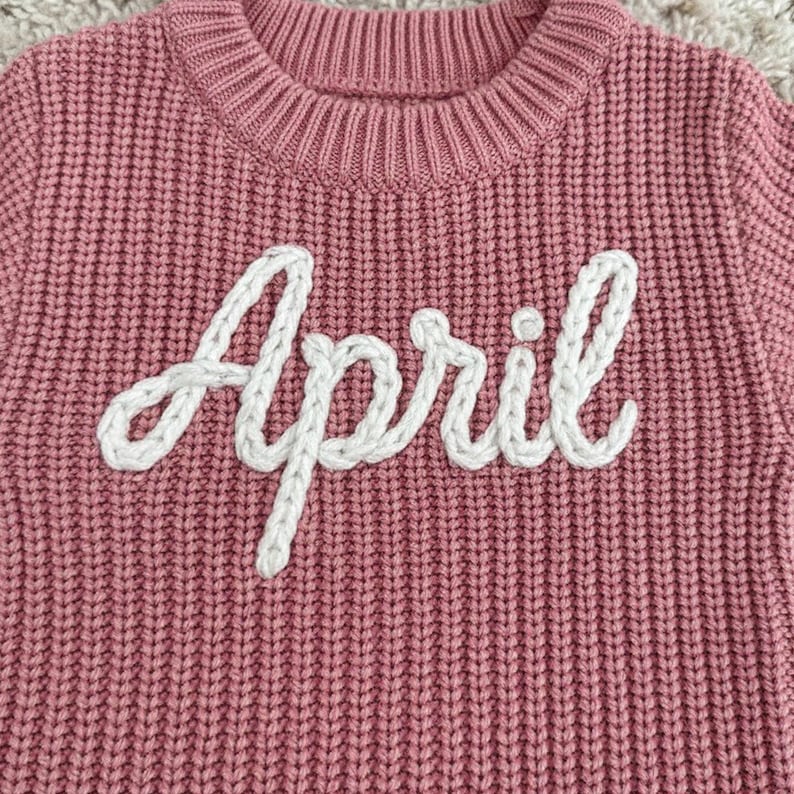 May include: A close-up of a pink knitted sweater with the word "April" embroidered in white. The sweater has a ribbed texture and a crew neck. The white embroidery is in a cursive font.