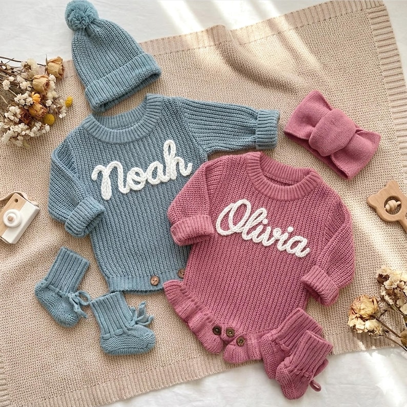 May include: A baby clothing set in shades of blue and pink, featuring a sweater with "Noah" in white script, a pink romper with "Olivia", a beanie, booties, and a headband. The items are arranged on a neutral-toned blanket.