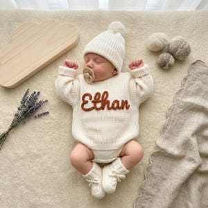 May include: A newborn baby wearing a cream-colored knit sweater with the name "Ethan" in brown letters, a matching beanie with a pom-pom, and booties. The baby is lying on a cream-colored surface with a wooden cloud-shaped object and lavender sprigs.