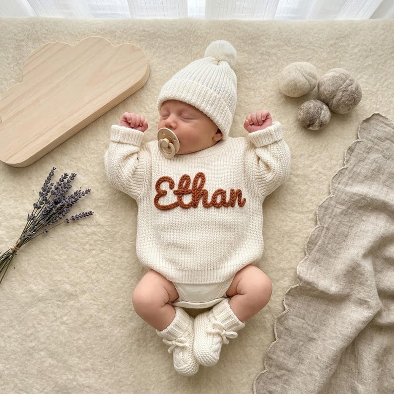 May include: A newborn baby wearing a cream-colored knit sweater with the name "Ethan" in brown letters, a matching beanie with a pom-pom, and booties. The baby is lying on a cream-colored surface with a wooden cloud-shaped object and lavender sprigs.