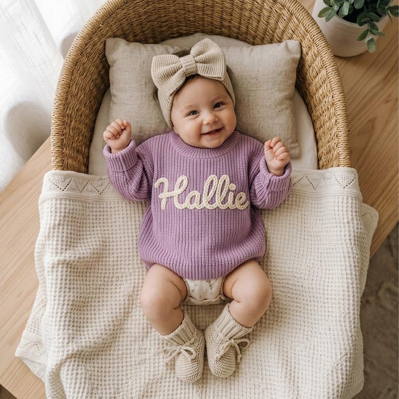 May include: A baby wearing a lavender long-sleeved knit jumper with the name Hallie embroidered in white script on the chest, a matching beige knit bow headband, and beige knit booties, lying on a white waffle-textured blanket.