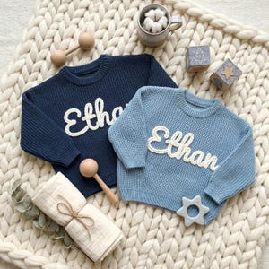 May include: Two knitted sweaters, one navy blue and one light blue, each with the name "Ethan" in white script. The sweaters are on a cream-colored, chunky knit blanket, with wooden toys and a swaddling blanket.