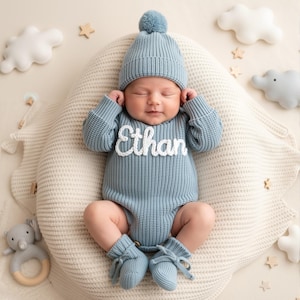 May include: A baby wearing a light blue knit outfit, including a hat with a pom-pom, a romper with the name "Ethan", and booties. The baby is lying on a cream-colored cushion, surrounded by decorative clouds, stars, and an elephant toy.