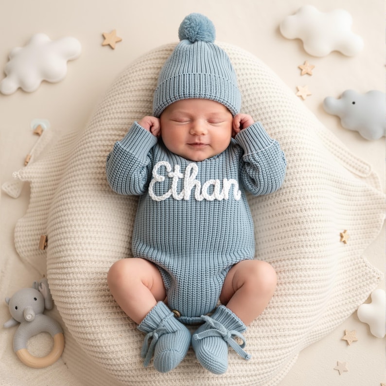 May include: A baby wearing a light blue knit outfit, including a hat with a pom-pom, a romper with the name "Ethan", and booties. The baby is lying on a cream-colored cushion, surrounded by decorative clouds, stars, and an elephant toy.