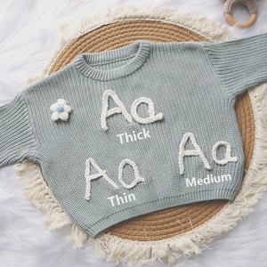 May include: A sage green knit sweater featuring three white embroidered letter A examples labeled Thick, Thin, and Medium, along with a small white flower appliqu&eacute; with a blue center. The sweater has a ribbed crew neckline and long sleeves.