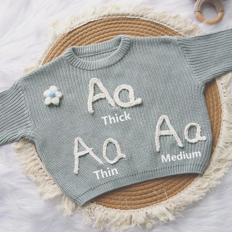 May include: A sage green knit sweater featuring three white embroidered letter A examples labeled Thick, Thin, and Medium, along with a small white flower appliqu&eacute; with a blue center. The sweater has a ribbed crew neckline and long sleeves.