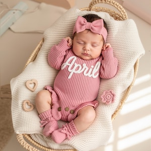 May include: A newborn baby wearing a pink knitted outfit with the name "April" in white script. The outfit includes a headband with a bow, a long-sleeved onesie, and matching socks. The baby is lying in a woven basket on a cream-colored blanket.