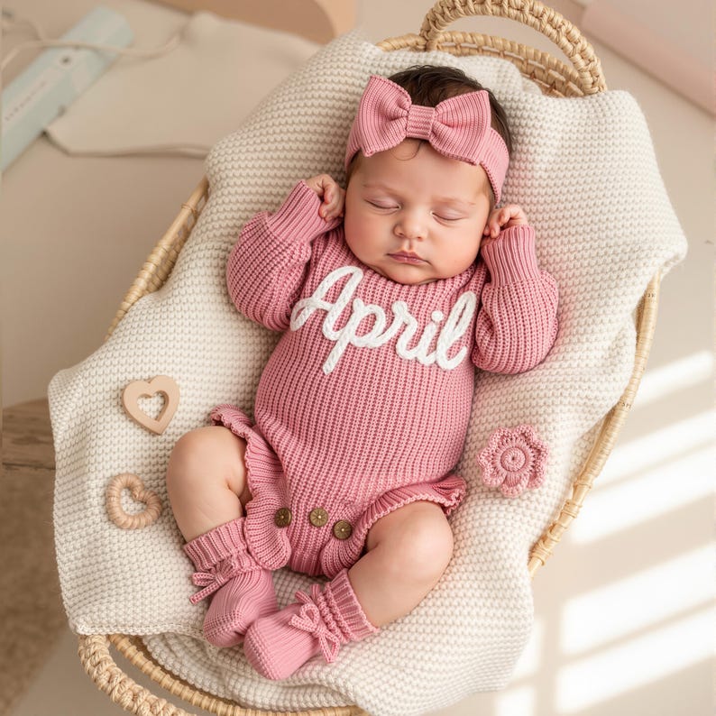 May include: A newborn baby wearing a pink knitted outfit with the name "April" in white script. The outfit includes a headband with a bow, a long-sleeved onesie, and matching socks. The baby is lying in a woven basket on a cream-colored blanket.