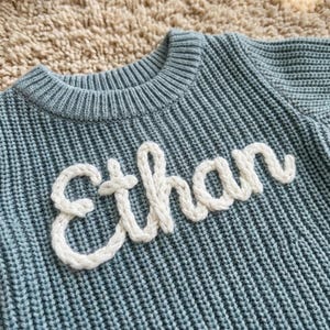 May include: A light blue knit sweater with the name "Ethan" in white, textured letters. The sweater has a ribbed crew neck and a textured knit pattern. The name is stitched onto the front of the sweater.