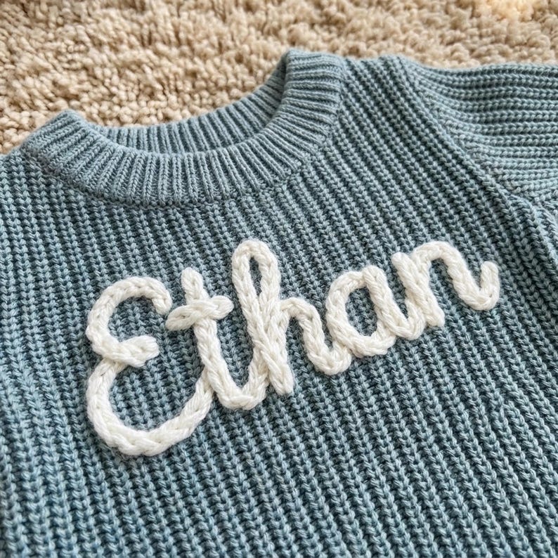 May include: A light blue knit sweater with the name "Ethan" in white, textured letters. The sweater has a ribbed crew neck and a textured knit pattern. The name is stitched onto the front of the sweater.