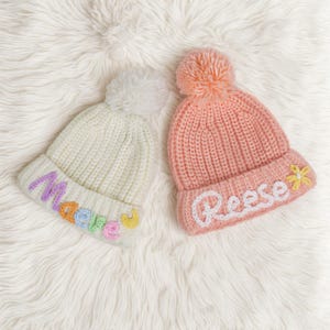 Pu&ograve; includere: Due berretti di lana con pon pon. Un berretto &egrave; bianco con il nome "Madelyn" ricamato in colori arcobaleno. L'altro berretto &egrave; rosa con il nome "Reese" ricamato in bianco.