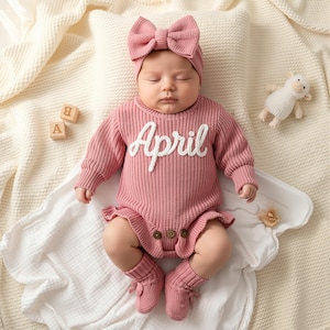 May include: A baby wearing a pink knitted outfit with the name "April" in white script. The outfit includes a bow headband, long-sleeved bodysuit, and socks. Wooden blocks with letters and a stuffed lamb are nearby.