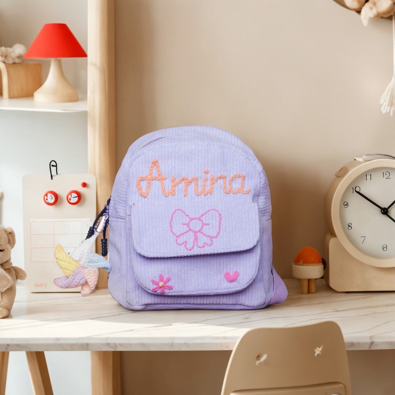 May include: A purple corduroy backpack with a pink bow and the name "Amina" embroidered on the front. The backpack has a small pocket on the front with a flower and heart design.