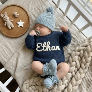 May include: A sleeping baby wearing a navy blue knit sweater with the name "Ethan" in white script, a light blue knit hat, and matching booties. A wooden tray with a stuffed bunny, a small bear, and a star is nearby.