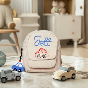 May include: A cream-colored corduroy backpack with the name "Jett" embroidered in blue thread. The backpack has a red and blue car design embroidered on the front flap.