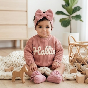 May include: A dusty rose knitted jumper featuring the name Hallie in white script lettering across the chest, paired with a matching knitted headband with a large bow and soft knitted booties, creating a coordinated set for an infant.
