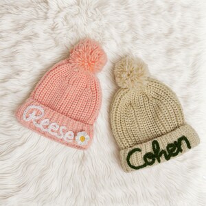 Pu&ograve; includere: Due berretti di lana con pon pon. Uno &egrave; rosa con un fiore bianco e il nome "Reese" ricamato. L'altro &egrave; beige con il nome "Cohen" ricamato in verde.