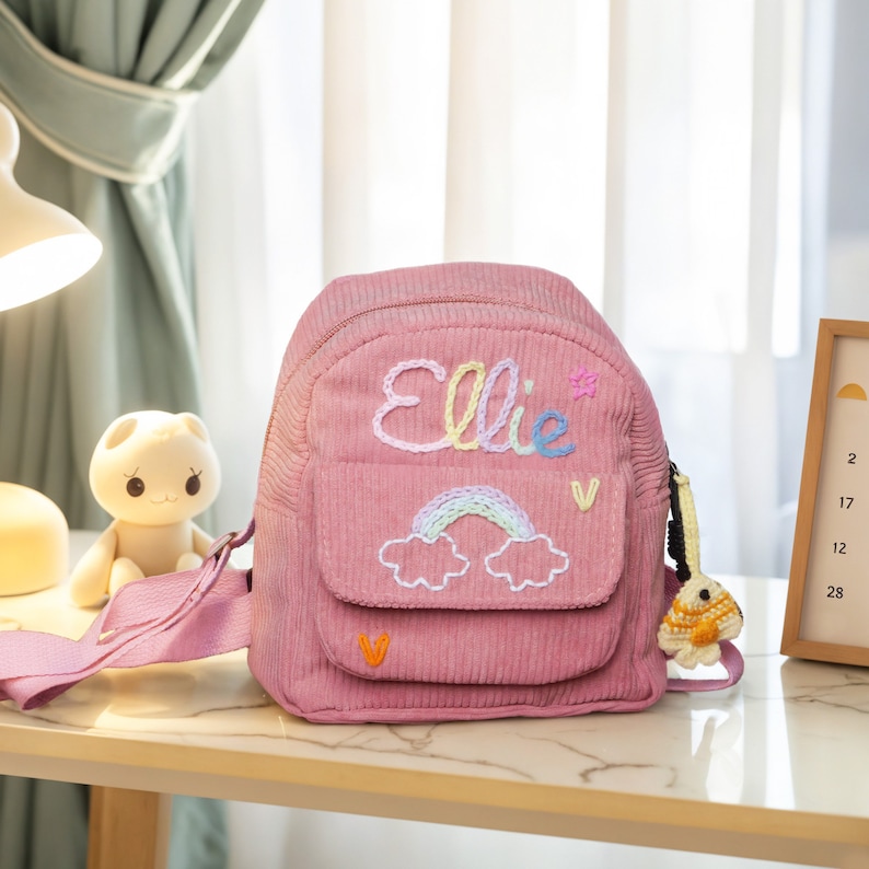 May include: Pink corduroy mini backpack with a rainbow and clouds embroidered on the front. The backpack has a zipper closure and a small pocket on the front. The name "Ellie" is embroidered on the front of the backpack in colorful thread.