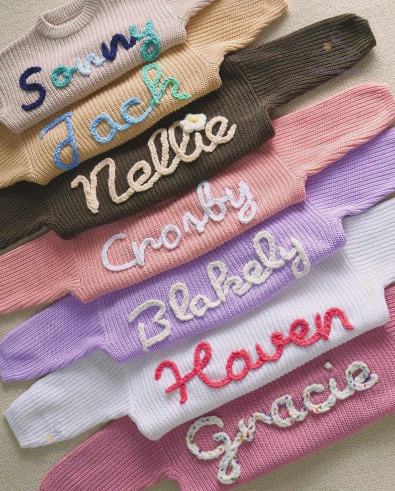 May include: A collection of long-sleeved knit sweaters in various colors including beige, brown, pink, lavender, white, and rose. Each sweater features a name embroidered in raised yarn lettering: Sonny, Jack, Nellie, Crosby, Blakely, Haven, and Gracie.