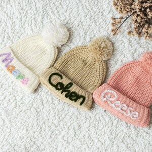 Pu&ograve; includere: Tre berretti di lana con pon pon. I berretti sono bianco, beige e rosa. Il berretto bianco ha un disegno arcobaleno e il testo "Madea". Il berretto beige ha il testo "Cohen" e il berretto rosa ha il testo "Reese".