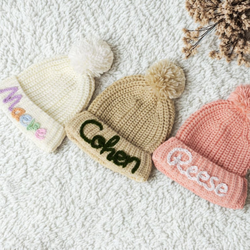 Pu&ograve; includere: Tre berretti di lana con pon pon. I berretti sono bianco, beige e rosa. Il berretto bianco ha un disegno arcobaleno e il testo "Madea". Il berretto beige ha il testo "Cohen" e il berretto rosa ha il testo "Reese".