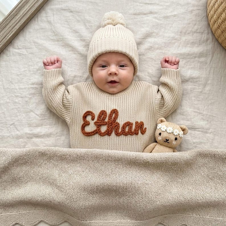 May include: A baby wearing a cream-colored knit sweater with the name "Ethan" in brown, a matching beanie with a pom-pom, and a beige blanket. A small teddy bear with a floral crown is next to the baby.
