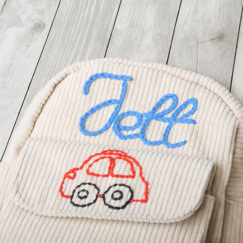 May include: A white corduroy backpack with the name "Jeff" embroidered in blue thread. The backpack also has a red car embroidered on the front flap.