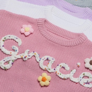 May include: A pink knit sweater featuring the word Gracie embroidered in white yarn with colorful flecks. The sweater is decorated with small, three-dimensional fabric flowers in pink, yellow, and white. Additional sweaters in white, lavender, and gray are stacked behind it.