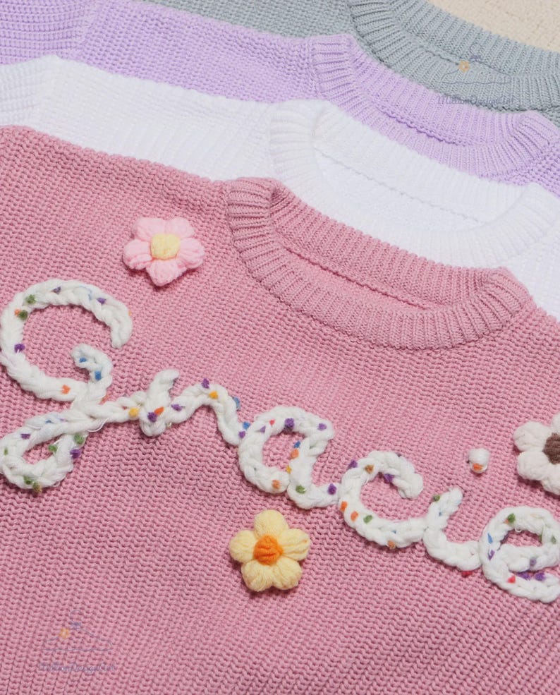 May include: A pink knit sweater featuring the word Gracie embroidered in white yarn with colorful flecks. The sweater is decorated with small, three-dimensional fabric flowers in pink, yellow, and white. Additional sweaters in white, lavender, and gray are stacked behind it.