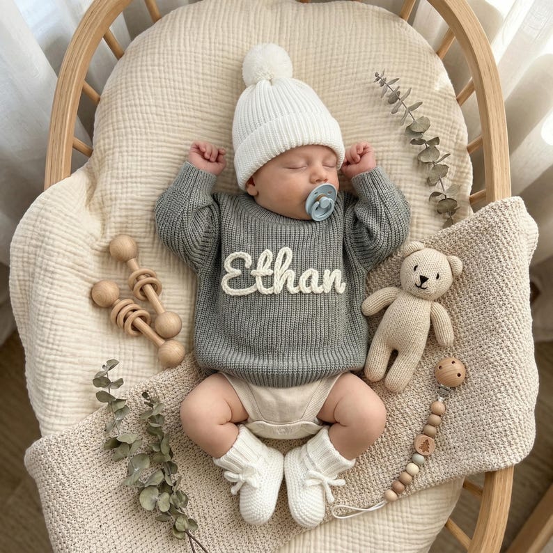 May include: A sleeping baby wearing a gray sweater with the name "Ethan" in white, a white beanie, and white booties. The baby is in a wooden cradle with a beige blanket, a teddy bear, and a wooden rattle.