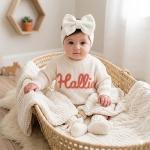 May include: A cream-coloured knit jumper featuring the name Hallie embroidered in a textured coral-coloured yarn across the chest, paired with a matching cream-coloured knit headband with a large bow and cream-coloured knit baby booties.