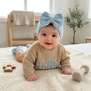 May include: A light blue knit headband with a large bow, a beige long-sleeved knit jumper with the name Hallie embroidered in light blue script, and matching light blue knit booties. The jumper features a ribbed texture and crew neckline.