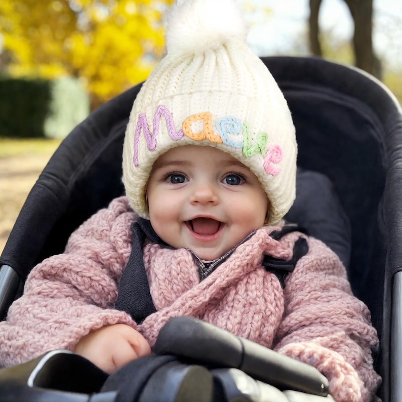 Pu&ograve; includere: Una bambina che indossa un cappello bianco di lana con un pon pon e il nome "Maeve" ricamato in lettere colorate. &Egrave; seduta in un passeggino nero e sorride.