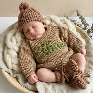 May include: A sleeping baby wearing a brown knit sweater with the name "Ethan" embroidered in green, matching knit booties, and a knit beanie with a pom-pom. The baby is lying in a wooden basket.