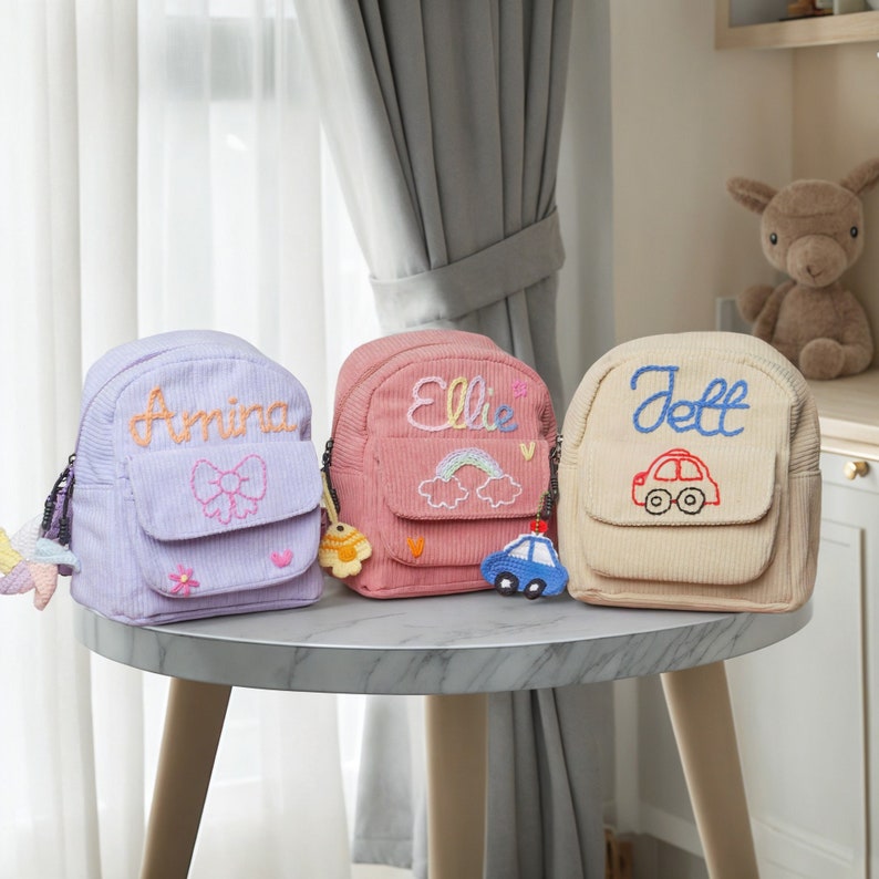 May include: Three small, personalized backpacks with embroidered names. The backpacks are in lavender, pink, and beige with different embroidered designs. The lavender backpack has a bow, the pink backpack has a rainbow and clouds, and the beige backpack has a red car.