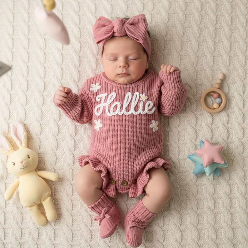 May include: A baby wearing a pink knit outfit with the name "Hallie" embroidered in white. The outfit includes a matching headband, socks, and a romper with floral accents. A plush bunny, wooden rattle, and star-shaped toy are nearby.