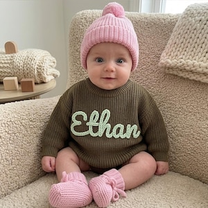 May include: A baby wearing a pink knit beanie with a matching pair of booties. The baby is also wearing a green knit romper with the name "Ethan" in light green script. The baby is sitting in a beige chair.