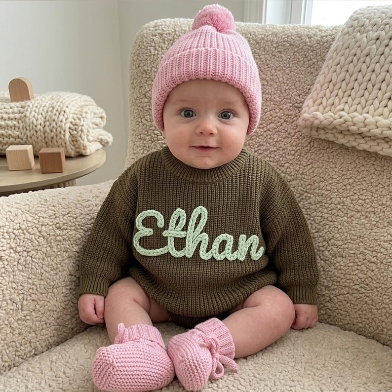 May include: A baby wearing a pink knit beanie with a matching pair of booties. The baby is also wearing a green knit romper with the name "Ethan" in light green script. The baby is sitting in a beige chair.