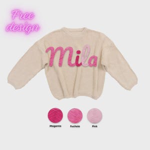 May include: A beige long-sleeve knit sweater featuring the name Mila in cursive script across the chest, with letters in varying shades of pink. Below the sweater are three circular yarn samples labeled Magenta, Fuchsia, and Pink.