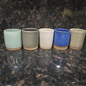 May include: Five handmade ceramic tumblers in various colors: light green, gray, speckled white, blue, and tan. Each tumbler has a cylindrical shape with a cork base. The tumblers are arranged in a row, showcasing their unique textures and hues.