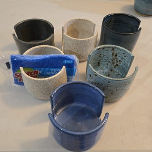 May include: A collection of handmade ceramic sponge holders in various colors, including blue, speckled beige, and dark gray. Each holder has a curved design with a slot for a sponge. A blue sponge is placed in one of the holders.