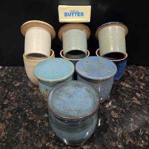 May include: A collection of ceramic butter keepers in various shades of blue, beige, and tan. The keepers have a cylindrical base and a matching lid. A stick of butter with the text "GRADE AA BUTTER" is in the background.