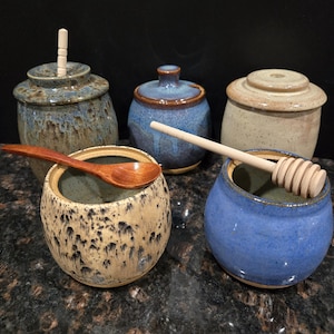 May include: Four ceramic honey pots with lids, each with a unique glaze. Two pots have wooden honey dippers, one has a wooden spoon, and one has a wooden stick.