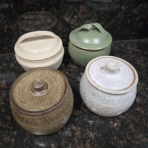 May include: Four ceramic jars with lids in various colors. One is beige with a handle, another is green with a looped handle, and two are brown and white with knob handles. The jars have textured surfaces.
