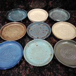 May include: Nine ceramic grater plates in various colors, including blue, beige, and brown. Each plate has a textured grating surface in the center. The plates are arranged in a grid pattern, showcasing their diverse hues and designs.