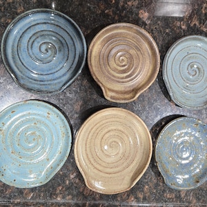 May include: Six handmade ceramic spoon rests in shades of blue and tan. Each rest features a spiral design and a small lip for holding utensils. The pottery has a rustic, speckled finish, perfect for kitchen decor.