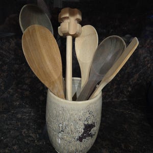 May include: A collection of wooden kitchen utensils, including spoons and a whisk, displayed in a speckled ceramic holder. The spoons vary in size and wood tone, creating a natural, rustic aesthetic. The holder is a neutral color with a textured surface.