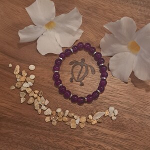 May include: Purple beaded bracelet with a silver turtle charm. The bracelet is on a wooden surface with white flowers and small white stones.