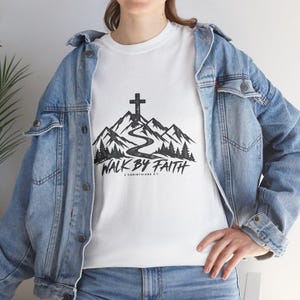 Puede incluir: Una camiseta blanca con un gráfico negro de una cordillera, un camino sinuoso, una cruz en la cima y un bosque. El texto dice WALK BY FAITH y 2 CORINTHIANS 5:7. La camiseta se lleva debajo de una chaqueta vaquera de color azul claro.