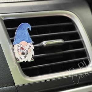 May include: A blue and white gnome holding a hockey stick and a hockey skate. The gnome is wearing a blue hat and has a white beard. The gnome is attached to a car air vent.
